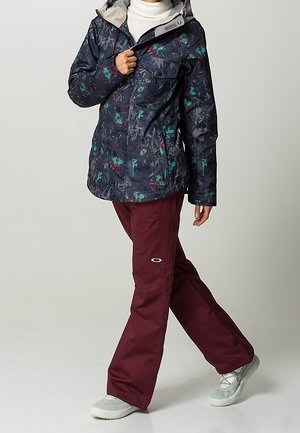 Waterdichte donkerblauwe jas met bloemenpatroon, grijze voering en met ritsen sluitende zakken; gecombineerd met bordeauxrode skibroek en grijze schoenen.