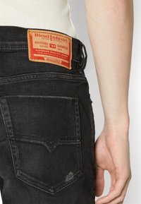 Svarta denimjeans med en texturerad finish, med ett läderpatch i röd och brun färg, två bakfickor och sömnadsdetaljer på bakstycket.