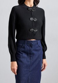 Svart ribbad topp med tre dekorerande knappar och långa ärmar, tillsammans med en mörk denimkjol. Texturerad tyg kontrasterar med slät denim.