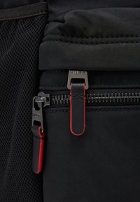 Borsa nera in tessuto con una superficie liscia; presenta una cerniera metallica con un tirazip con il marchio e un accento rosso, e una tasca in rete sul lato.