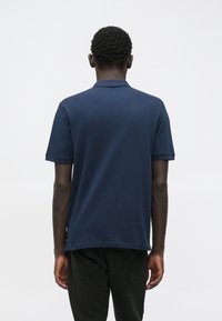 Homme debout, dos tourné, portant un polo bleu marine à manches courtes et un pantalon noir sur un fond clair uni.