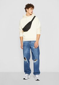 Sac en bandoulière noir porté sur un sweatshirt crème ; modèle en jeans déchirés bleu clair et baskets blanches avec accents gris.