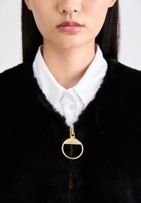 Schwarze Kunstpelzjacke mit einem goldenen Reißverschluss, der einen runden Ring und ein Markenlogo aufweist, getragen über einem weißen Hemd mit Kragen.