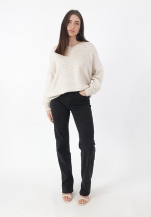 Lichtbeige gebreide trui met een V-hals, gecombineerd met zwarte rechte jeans. Het model draagt tan open-teen sandalen. Eenvoudige, casual outfit.