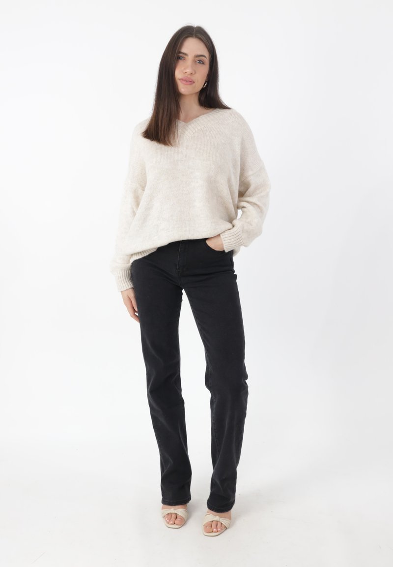 Maglione a maglia beige chiaro con scollo a V, abbinato a jeans neri a gamba dritta. Il modello indossa sandali aperti color carne. Outfit semplice e casual.