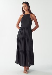 Žena v bezramínkových černých maxi šatech s volány a béžových sandálech, stojící s rukama za zády na bílém pozadí.