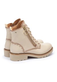 Botas de tobillo de cuero beige con cremallera lateral, herrajes dorados, acentos de tachuelas y suela de goma texturizada en tono marrón.