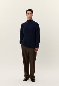 Moški stoji z rokami v žepih, oblečen v temno modro pleteno jopico, rjave hlače in črne čevlje, na ravni svetli podlagi.