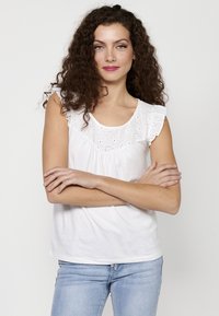Top senza maniche in cotone bianco con rifiniture arricciate sulle spalle e dettagli in pizzo lungo il collo. Abbinato a jeans in denim blu chiaro.