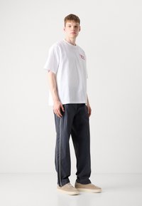 Witte katoenen t-shirt met rood logo, gecombineerd met marineblauwe joggingbroek met witte zijstrepen. Beige sneakers maken de outfit compleet.