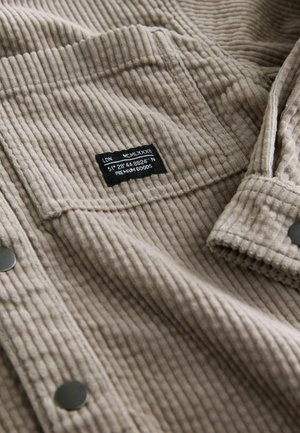 Beige kordfløjl med lodrette striber, prydet med en sort label med tekst og sølvfarvede trykknapper på forsiden. Strukturdetalje på overfladen.
