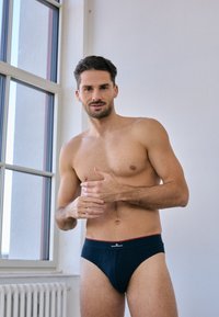 Marineblaue Baumwollunterhosen mit einem roten elastischen Bund und Logodetail, die eine enge Passform und glatte Textur bieten, präsentiert von einem Mann.