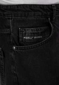 Jeans negros holgados de denim, con un bolsillo trasero que tiene una etiqueta visible "REELL BAGGY" y un botón metálico como detalle.