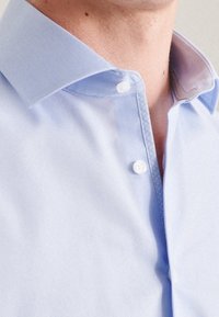 Primo piano di una persona che indossa una camicia di jeans leggera, con il primo bottone sbottonato, che mostra il colletto e parte del collo.