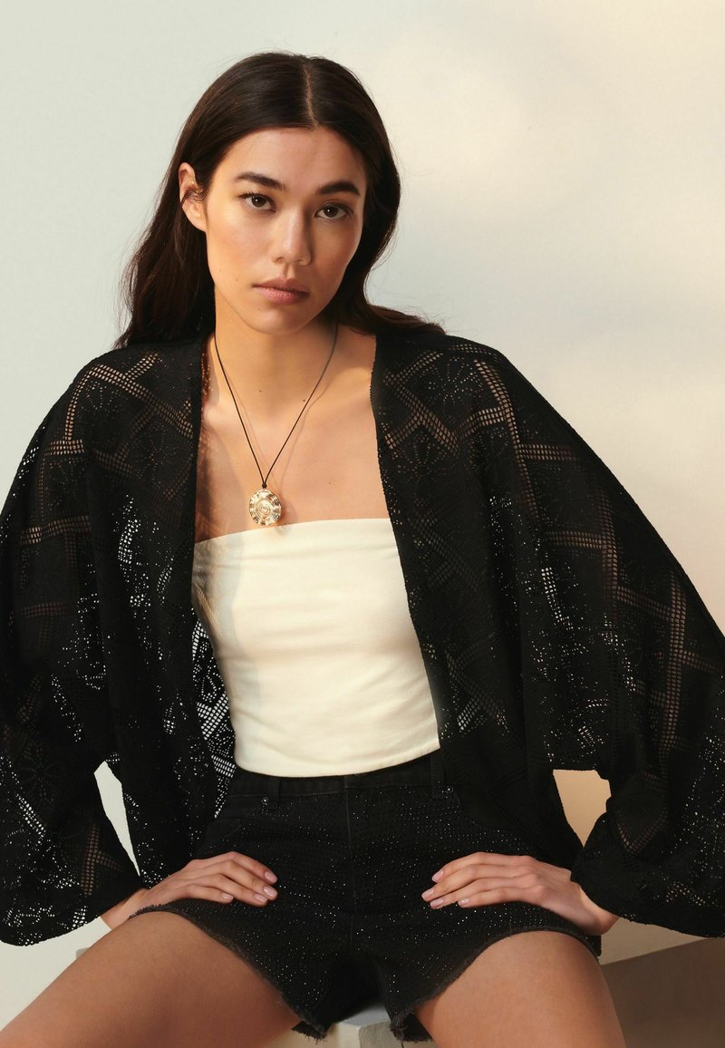 Svart spetsskimono med intrikata geometriska mönster, lager på en vit utan ärm topp, tillsammans med svarta texturerade shorts.