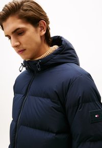 Giacca puffer blu navy con design trapuntato, chiusura con zip, cappuccio regolabile e patch con logo sulla manica sinistra. Collo di colore chiaro.