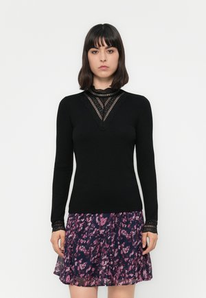 Woman wearing a black long-sleeve top with lace trim and a purple patterned skirt, standing against a plain white background.