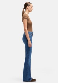 Dzwony z niebieskiego denimu o wysokim stanie, z klasycznym pięciokieszeniowym krojem i lekkim przetarciem, zestawione z dopasowanym brązowym topem.