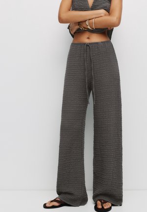 Femme portant un pantalon ample texturé gris foncé avec cordon, un top court sans manches, des bracelets dorés et des sandales tongs foncées, bras croisés.