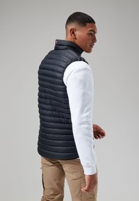 Marineblauw gewatteerd vest van lichtgewicht materiaal, voorzien van een rits en een hoge kraag, gedragen over een wit longsleeve shirt.