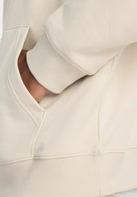 Felpa beige con tasca a marsupio, maniche a costine e texture liscia. Il tessuto ha una leggera lucentezza e un design dal taglio rilassato.