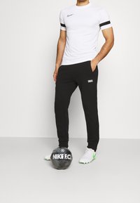 Valkoinen lyhythihainen Nike-paita mustilla yksityiskohdilla, yhdistettynä mustiin verkkareihin, joissa on logopatchi, sekä mustaan jalkapalloon.