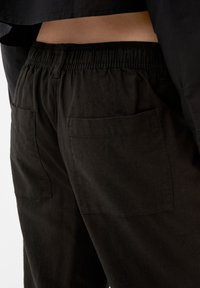 Pantalon en coton noir avec une taille élastique et deux poches arrière. La texture du tissu est douce avec un aspect légèrement usé.
