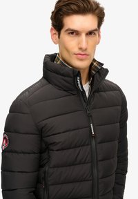 Giubbotto invernale nero con texture trapuntata, colletto alto e zip frontale. Include un logo sulla manica sinistra e un interno in tessuto a quadri.