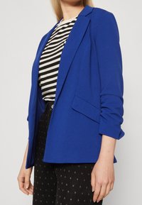 Blazer blu dalla vestibilità su misura, con spalline e design aperto. Indossato sopra una maglietta a righe bianche e nere, abbinato a pantaloni fantasia.