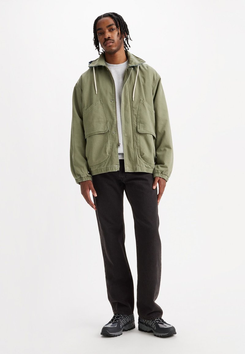 Olijfgroene lichtgewicht jas met twee grote voorzakken, een capuchon met trekkoord en elastische manchetten, gecombineerd met donkere jeans en zwarte schoenen.