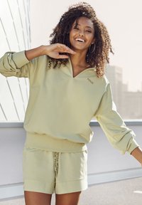 Light olive green sweatshirt with a deep V-neck and wide sleeves, paired with matching shorts featuring a drawstring and logo detailing.