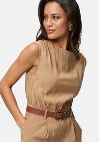 Robe beige élégante sans manches avec une taille ajustée, complétée par une ceinture marron avec une boucle ronde et un détail texturé.