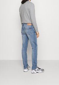 Jasnoniebieskie dżinsy denim o wąskim kroju, z skórzaną naszywką na talii, połączone z szarą koszulą z długim rękawem i białymi trampkami.