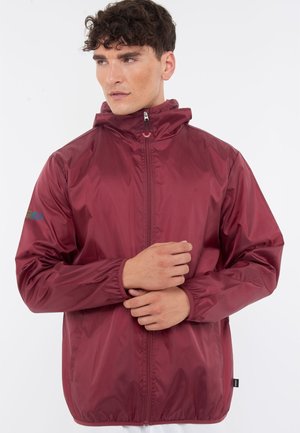 Jeune homme portant une veste coupe-vent bordeaux zippée avec des poignets élastiques, debout devant un fond blanc uni.