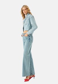 Chaqueta de mezclilla azul claro recortada con un patrón texturizado, combinada con unos vaqueros acampanados a juego. Unos zapatos de tacón rojos completan el conjunto.