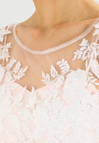 Vestido rosa claro con un escote transparente, con bordados de encaje blanco y acentos de cuentas, que presenta intrincados patrones florales.