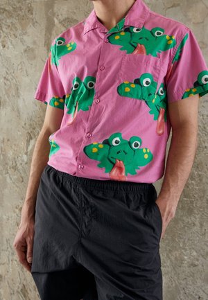 Uomo che indossa una camicia rosa a maniche corte con grandi facce di rana verdi che tirano fuori la lingua, abbinata a pantaloni neri.