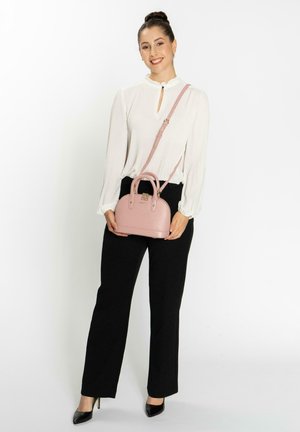 Light pink handbag with a textured finish, rounded top, double handles, and an adjustable shoulder strap. Worn with black pants and a white blouse.