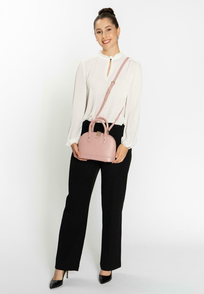 Bolso de mano rosa claro con un acabado texturizado, parte superior redondeada, doble asa y una correa de hombro ajustable. Combinado con pantalones negros y una blusa blanca.