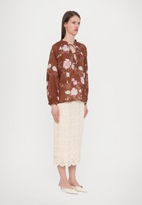 Bruine bloemenblouse met een keyhole-decolleté, gecombineerd met een witte gehaakte midi-jurk met een geschulpt zoom, en witte puntige muiltjes.