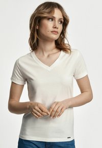 Biała koszulka z dekoltem w serek i krótkimi, podwiniętymi rękawami. Miękka tkanina, dopasowany krój oraz mały detal z logo na dole.