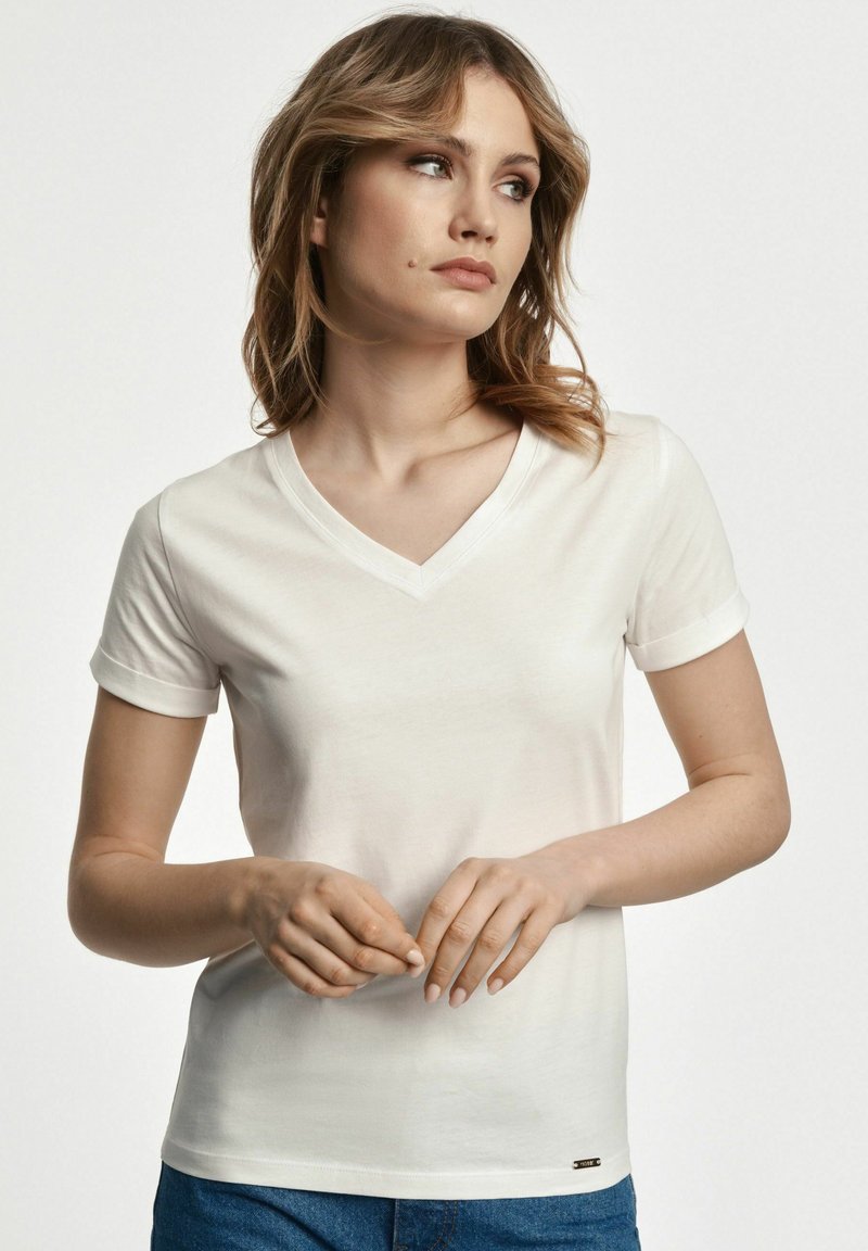 Biała koszulka z dekoltem w serek i krótkimi, podwiniętymi rękawami. Miękka tkanina, dopasowany krój oraz mały detal z logo na dole.