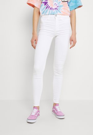 Personne portant un jean skinny blanc, des baskets roses et blanches, et un t-shirt coloré tie-dye, debout sur un fond blanc uni.