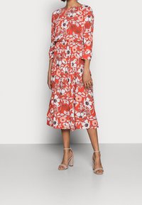 Robe midi florale d'un rouge vif avec des manches trois quarts, une taille cintrée et des motifs de fleurs blanches. Associée à des sandales à talons blocs nude.