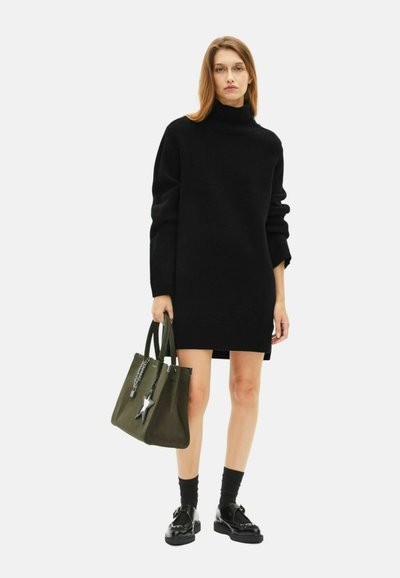 Schwarzer overgroßer Rollkragenpullover mit gerippten Bündchen, kombiniert mit einer grünen Tragetasche mit Sternakzent und schwarzen Schnürschuhen.