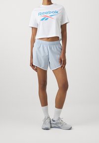 Bílý cropped tričko s modrým a červeným logem "Reebok", v kombinaci se světle modrými šortkami s bílým lemem. Model má na sobě šedé tenisky a bílé ponožky.