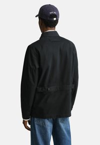 Mann, der eine schwarze Knopfleiste mit verstellbarem Rückengurt und eine marineblaue Mütze trägt, von hinten vor einem schlichten Hintergrund gezeigt.