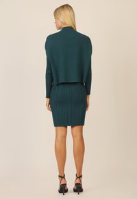 Türkiser gestrickter Pullover mit geripptem Textur, der über einem dazu passenden eng anliegenden gerippten Kleid hängt, das einen geraden Saum und lange Ärmel hat. Schwarze Riemchen-Heels.