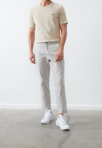 Homme portant un t-shirt beige, un pantalon gris clair et des baskets blanches, debout sur un sol en béton gris devant un mur blanc.