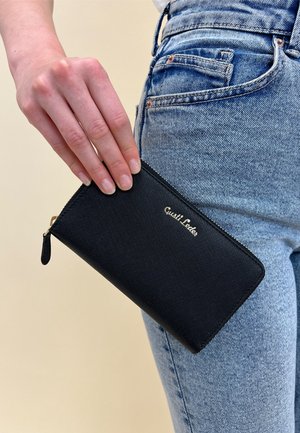 Black zip wallet made of textured material, featuring gold logo, rectangular shape, and a small zip pull tab; held near denim jeans.